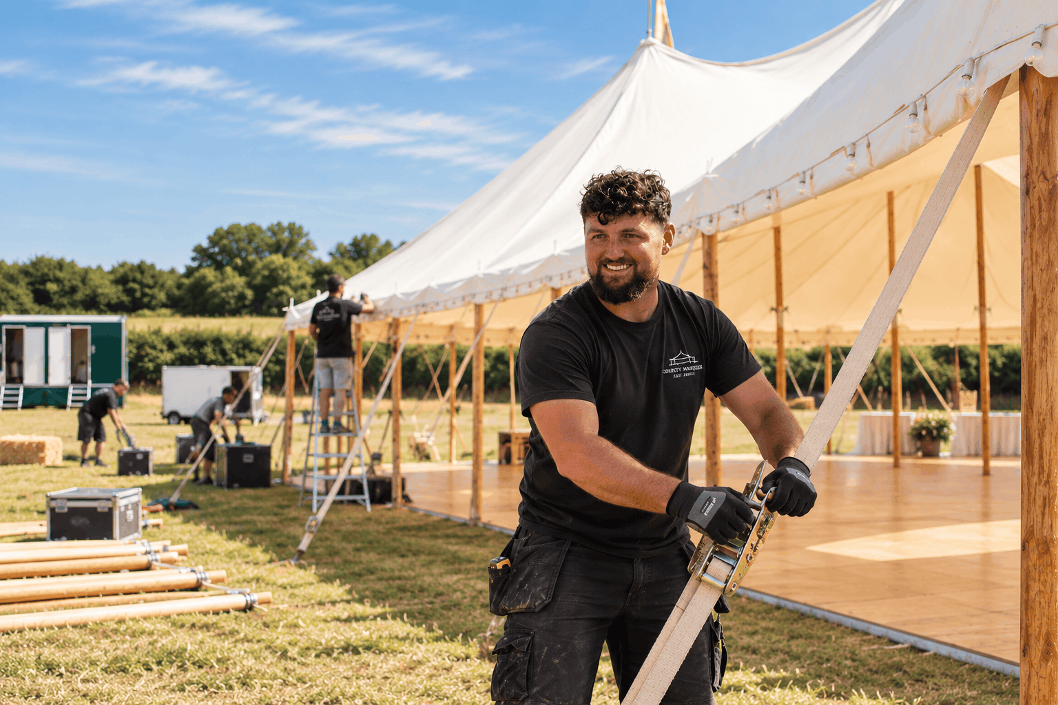 Man conducts marquee installation in field with other men in background installing same marquee